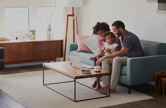 A family with a mom, dad, and toddler sitting on a living room couch and looking at a laptop on the coffee table